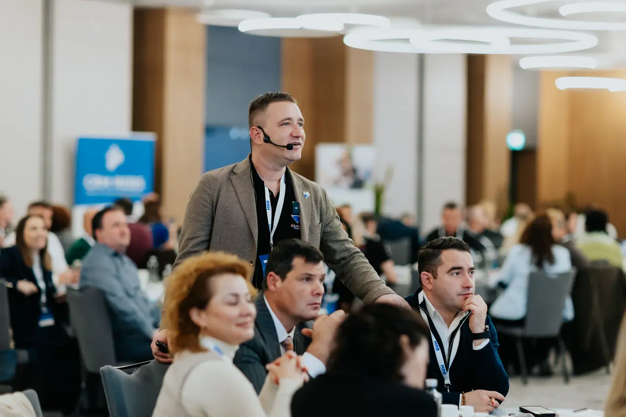 Moderator cu microfon headset vorbind printre mesele participanților, MRB 2025